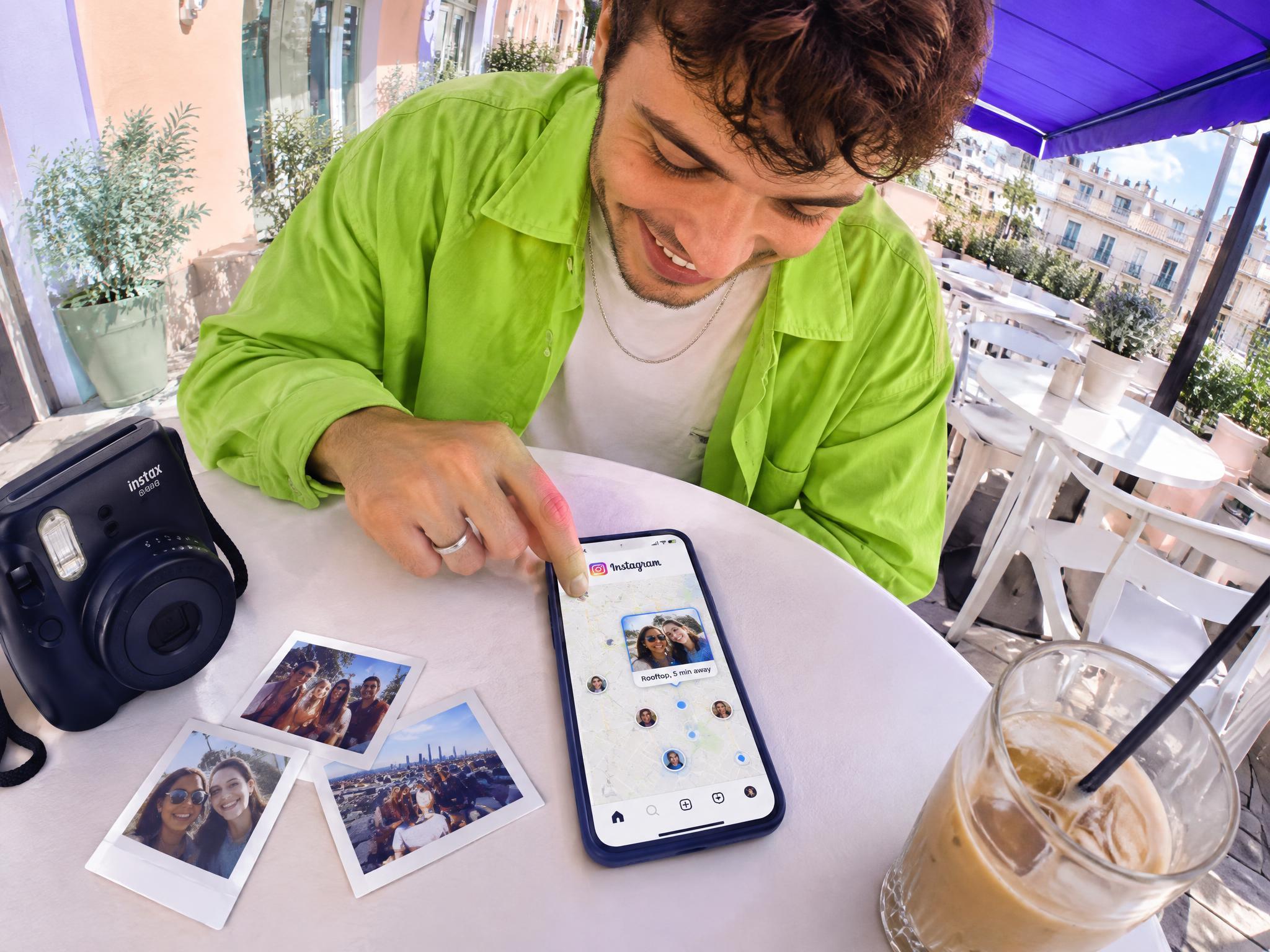 Young man at a café table using the Instagram Map feature on his phone, next to an instant camera and polaroid photos