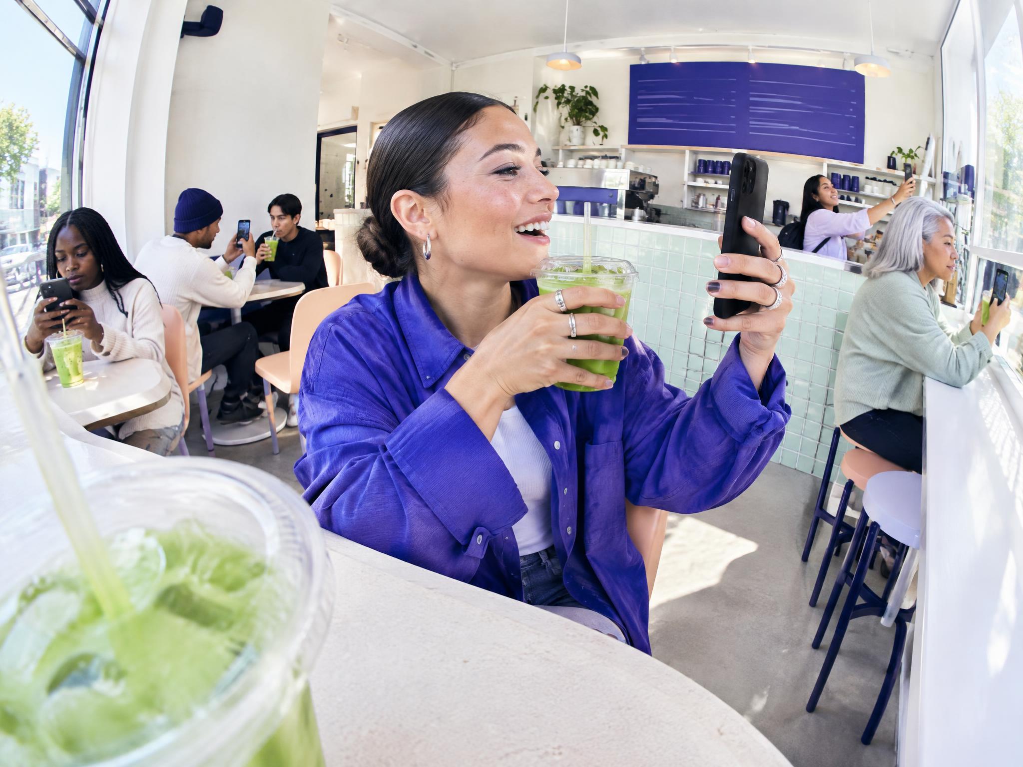 Woman in a café filming herself with a smartphone while holding a green drink