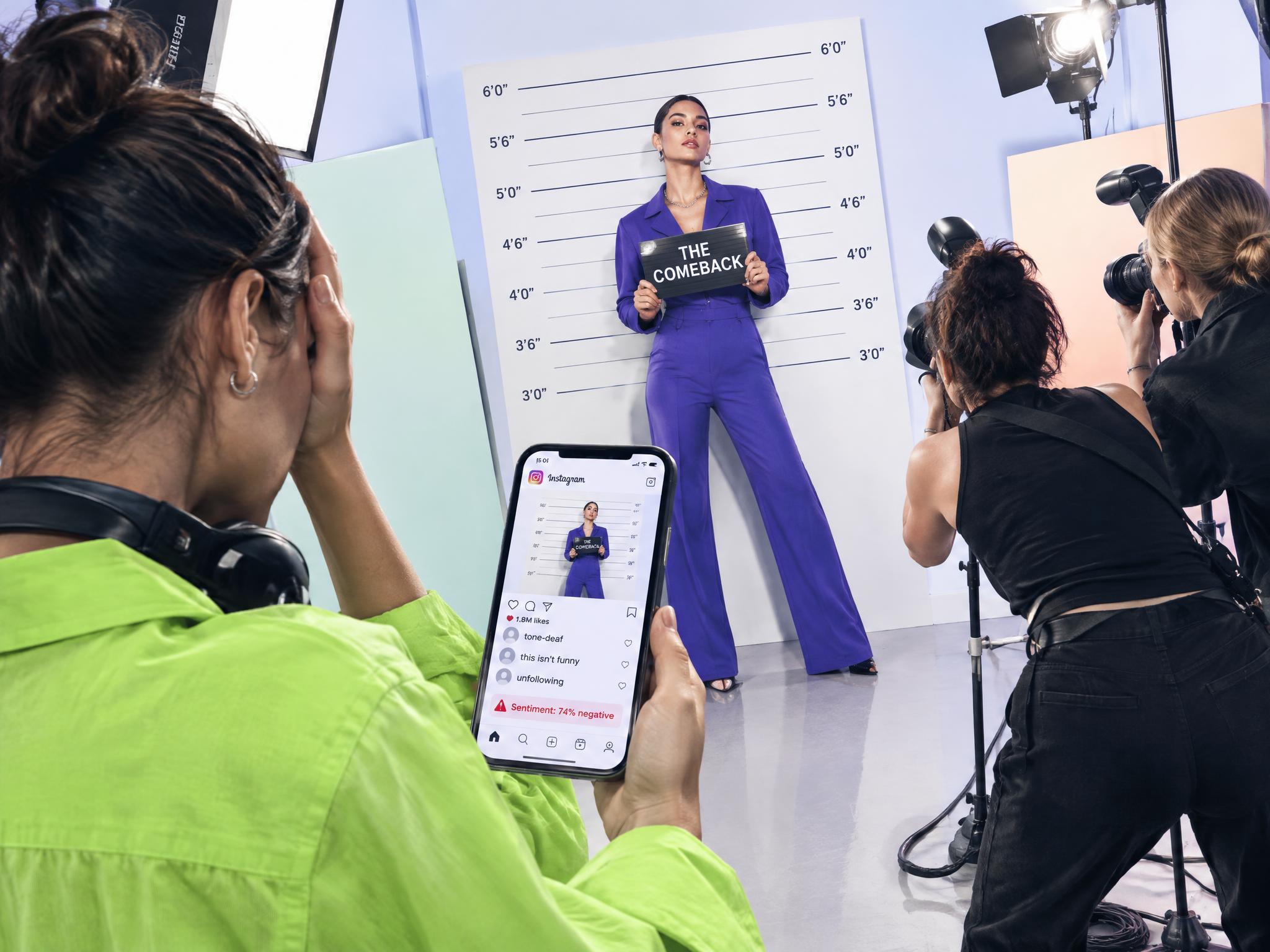 Woman holding a "The Comeback" sign at a mugshot-style photo shoot while negative comments show on a phone