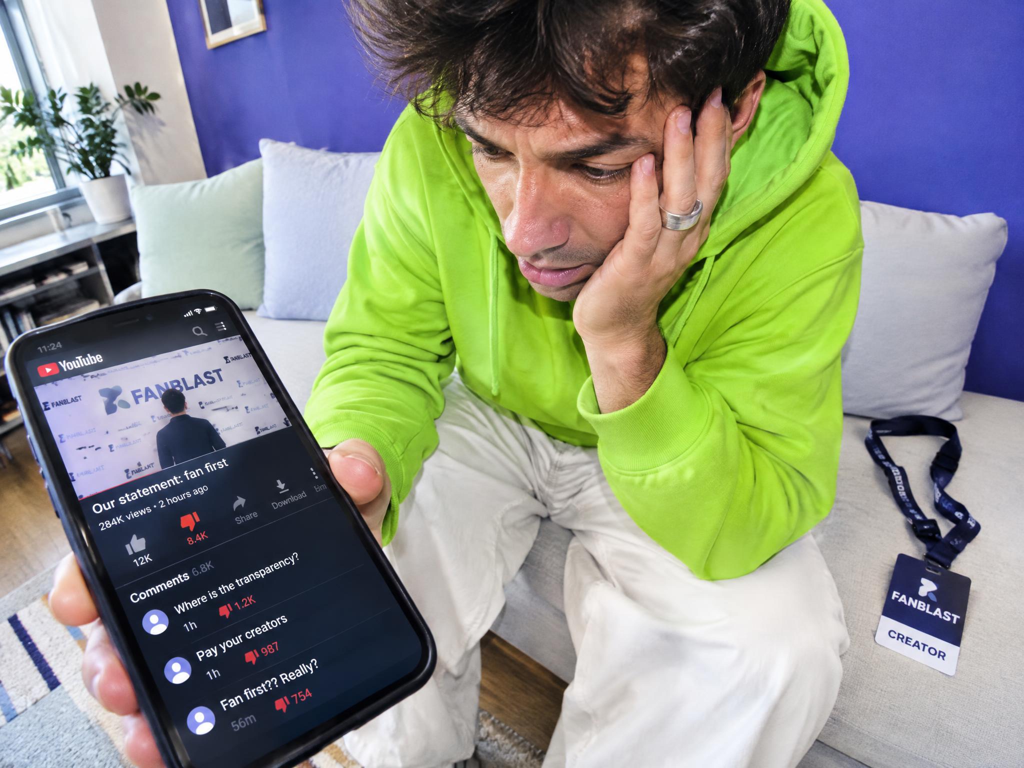 Worried man on a sofa holding a phone showing a Fanblast YouTube statement with critical comments