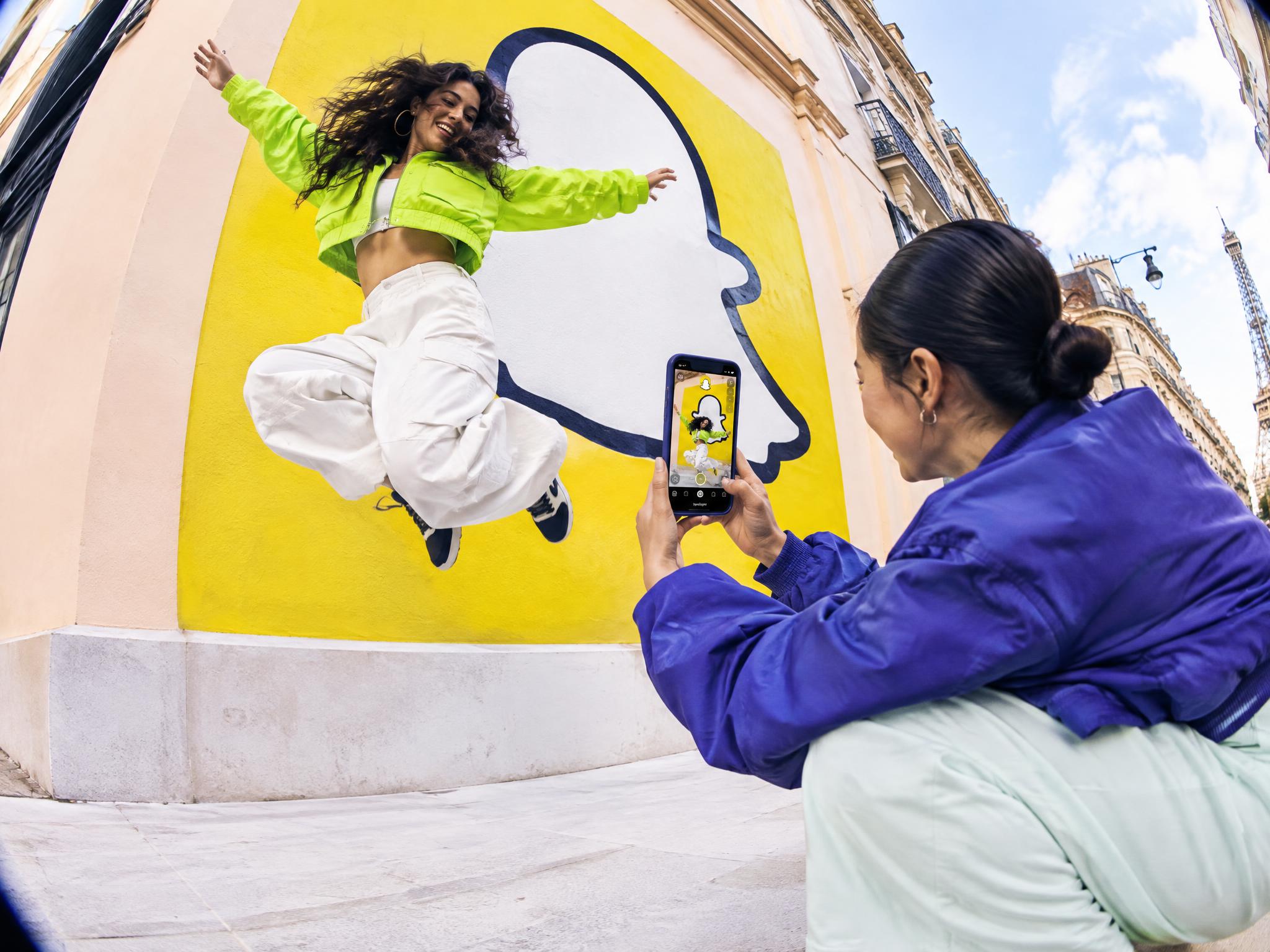 Woman photographing a friend jumping in front of a Snapchat logo mural in Paris