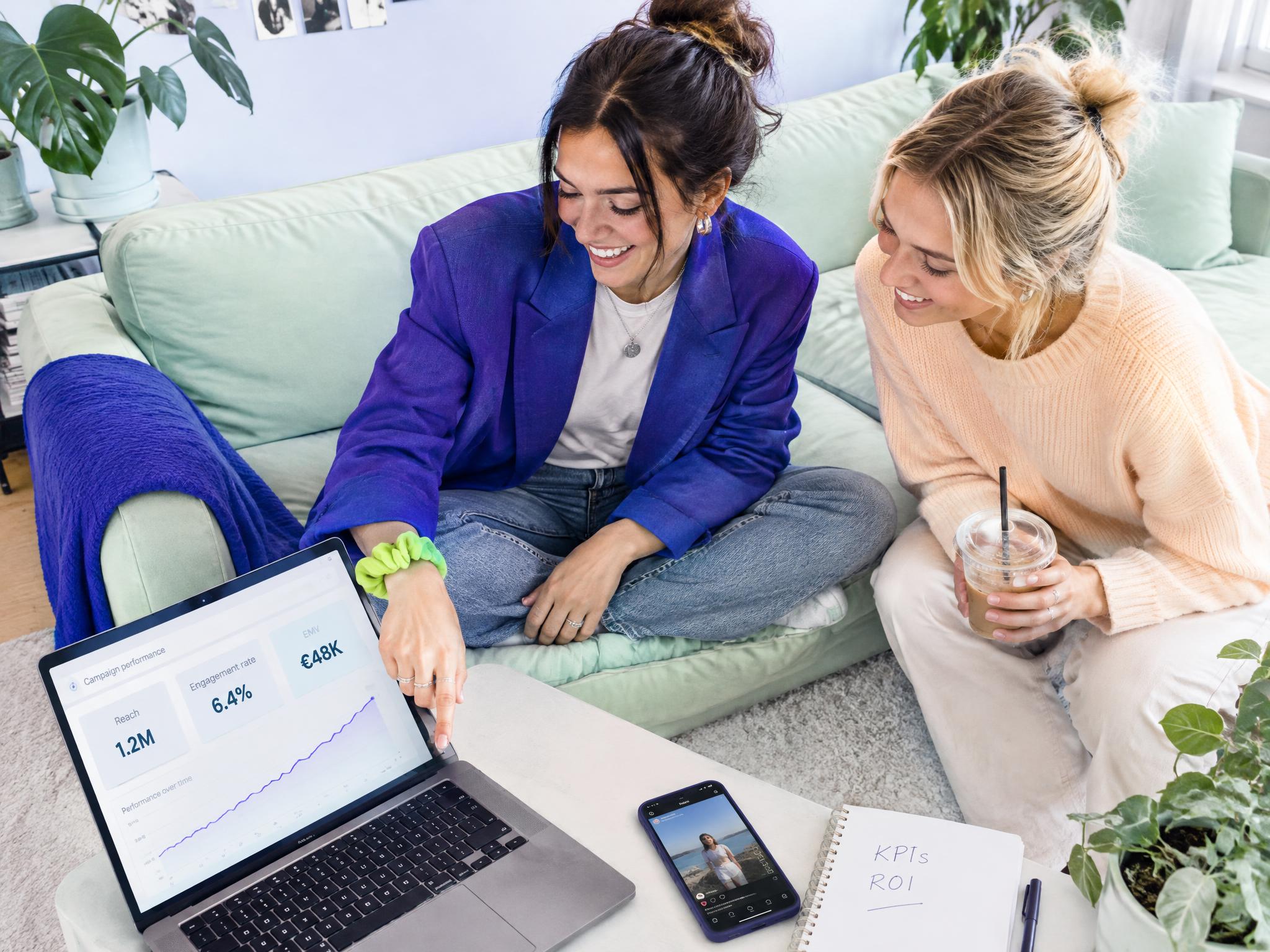 Two women on a sofa review a campaign performance dashboard showing reach, engagement rate and EMV on a laptop